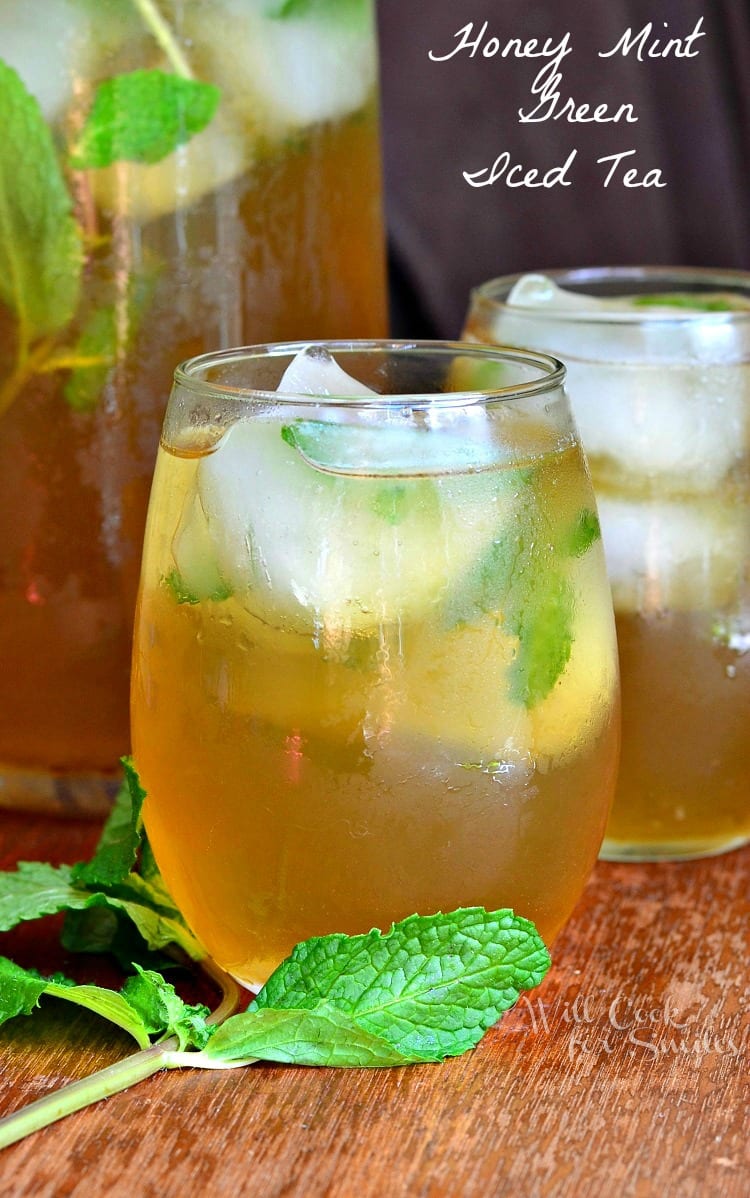 2 glasses of brewed honey mint green iced tea on a wooden table with mint laying on the table to the left of the glasses and a pitcher filled with more tea in the background to the left of glasses.