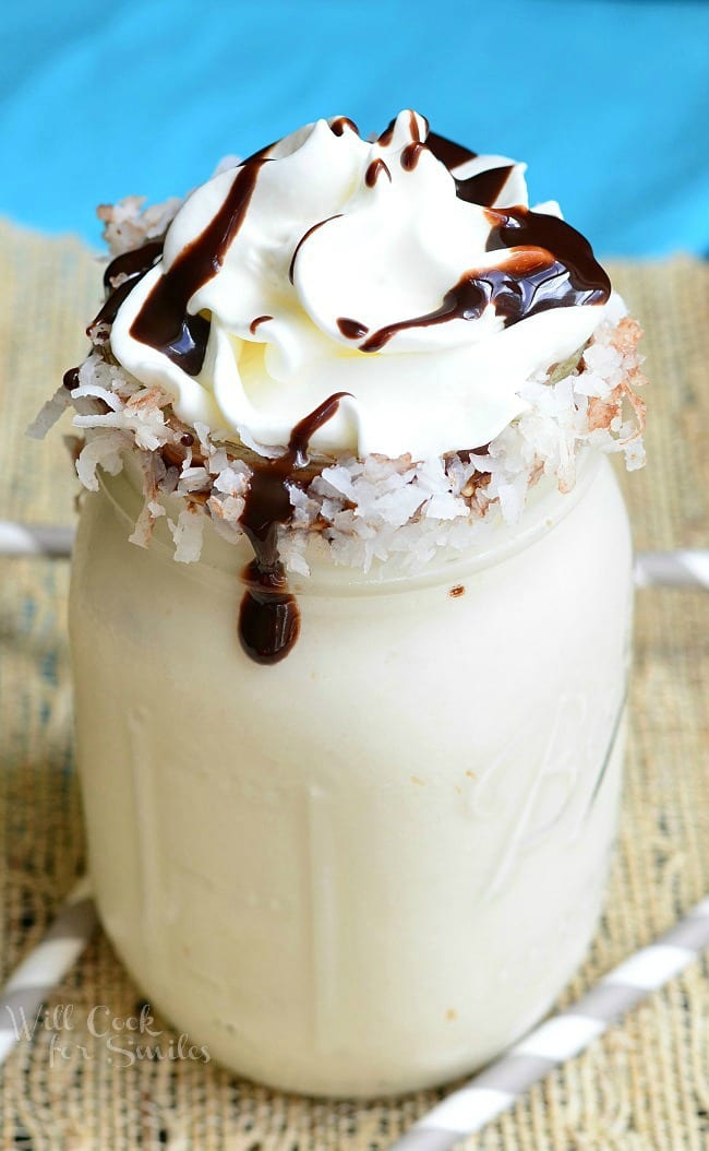Milkshake in a mason jar with coconut around the rim and whip cream on top with a chocolate sauce drizzle over it