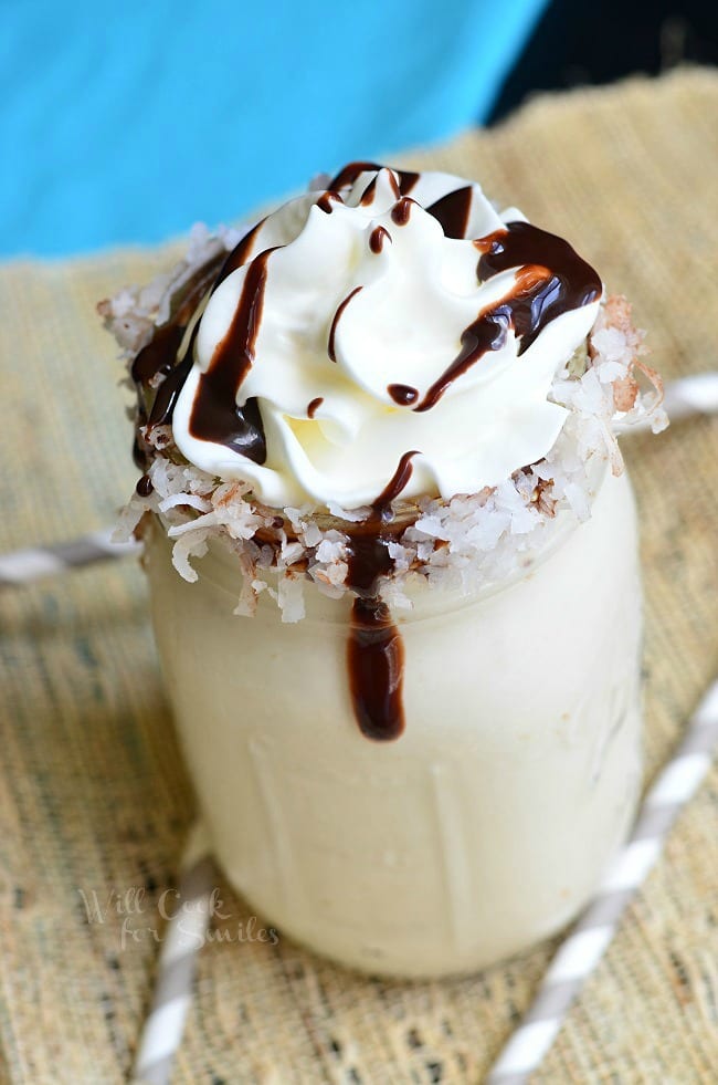 view from above of mason jar filled with lightened almond joy milkshake on a tan placemat with 3 straw around the base of the jar and a blue cloth in the background topped with whipped cream and chocolate syrup