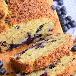 view from above of loaf of toasted coconut blueberry bread with the first 3 slices stacked in front of the rest of the loaf on a wooden cutting board with blueberries scattered around on table.