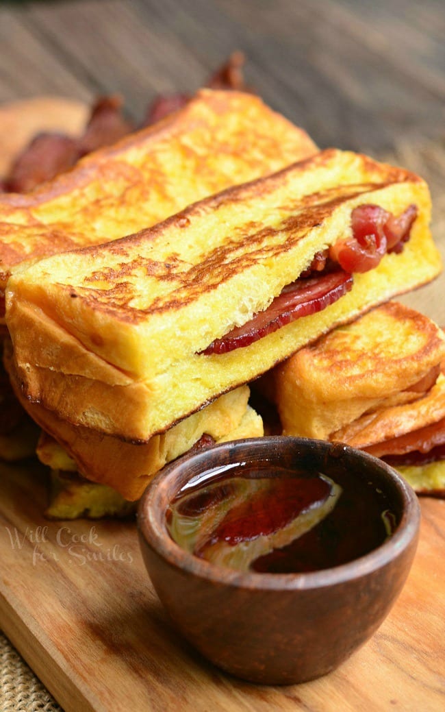 Cut bacon stuffed brioche french toast sticks stacked on a wooden cutting board with a small bowl of syrup