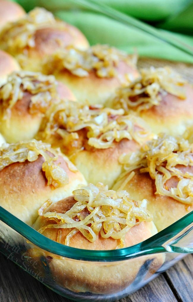 close up view of glass baking pan filled with caramelized onion dinner rolls topped with onions on a wooden table with a green cloth behind