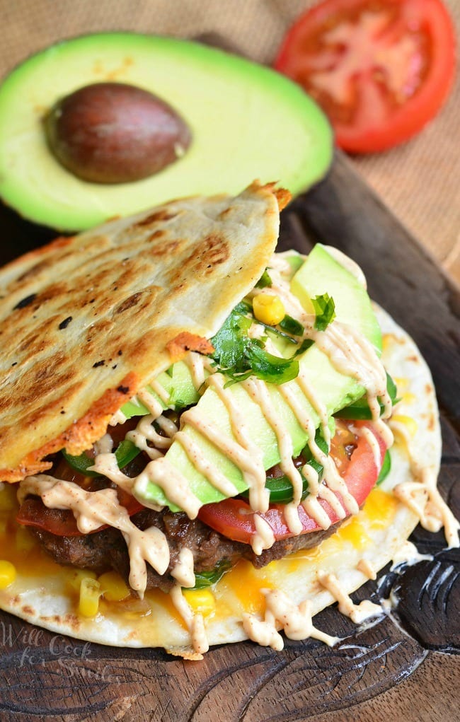 burger sandwiched between tortillas with avocado, tomato, and home made sauce and a half of avocado and tomato in the background
