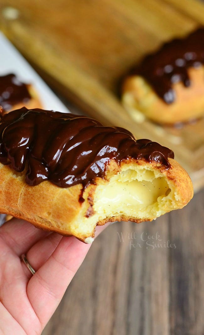 hand holding up one eclair with a bite in it above wooden table and more eclairs on a wooden cutting board