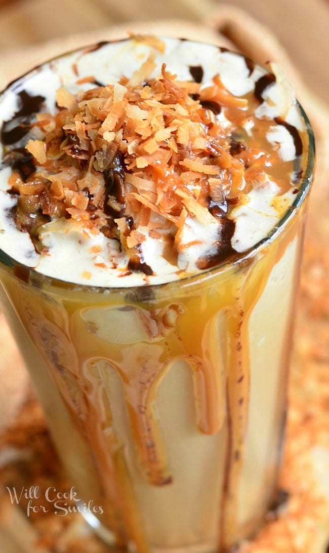 Samoas Frozen Coffee in a glass cup with whip cream, toasted coconut, chocolate sauce, and caramel on top. Caramel and chocolate sauce pouring down the outside of the glass