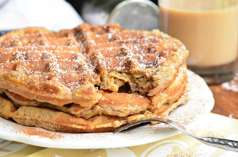 Side view of three Cinnamon Peach Oat Waffles stacked on top of each other served on a white plate. Waffles are sprinkled with cinnamon and powder sugar. A bite or piece of the top waffle is gone and the fork is set on the edge of the plate.