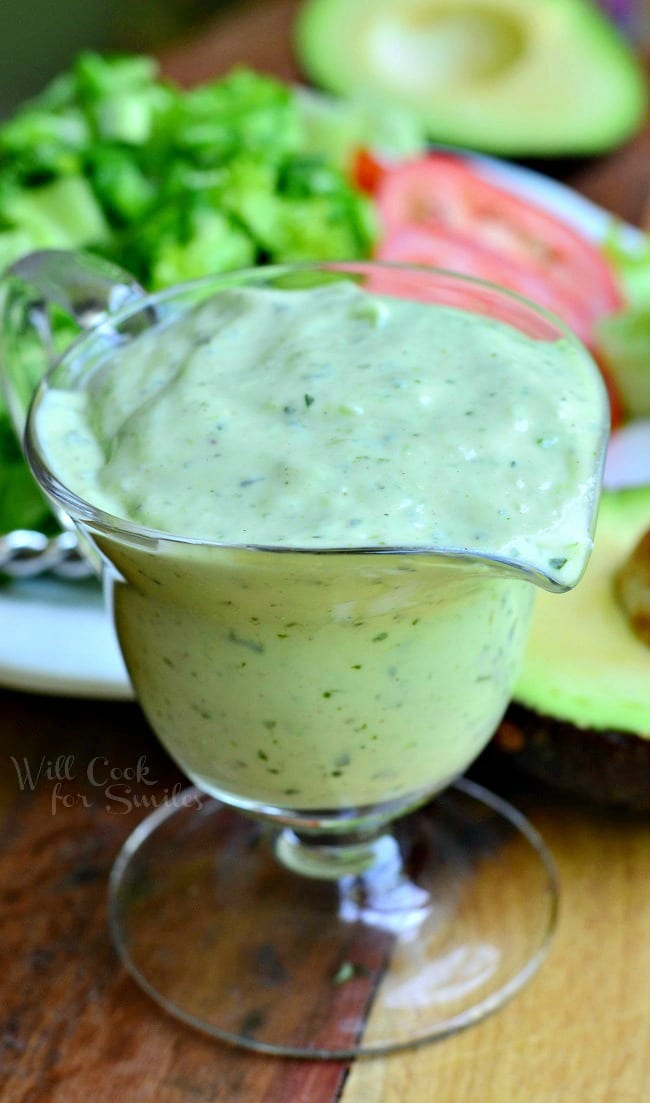 Sauce boat filled with creamy avocado dressing on a wood table with a salad on a white plate in the background