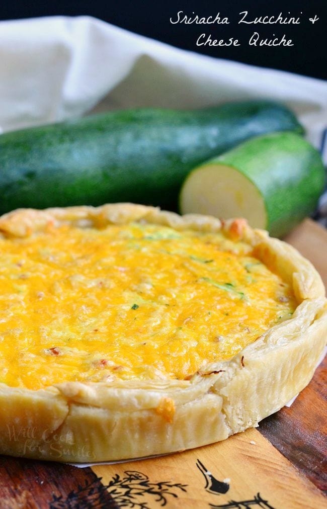 A whole Sriracha Zucchini and Cheese Quiche is laying on a wood board. The outside crust appears flaky and the top is covered with cheese. There are zucchinis in the background of this photo..