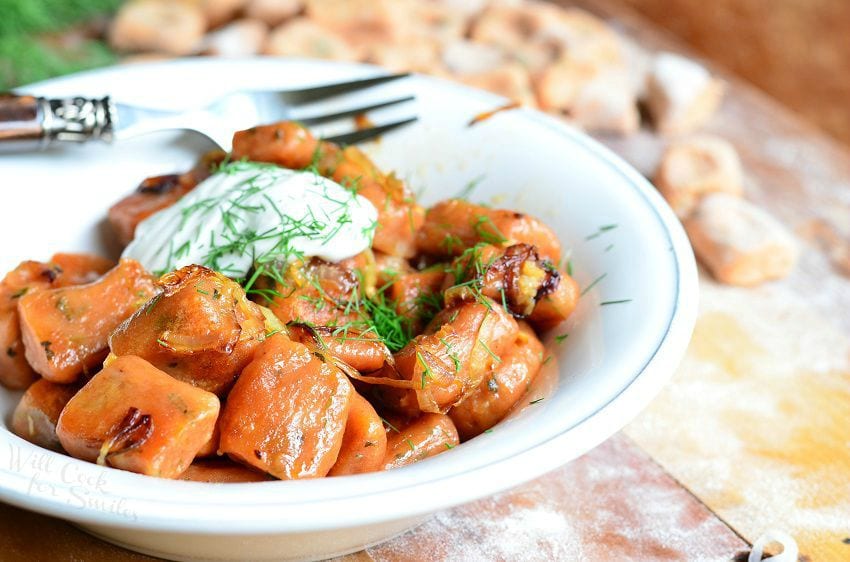 vertical photo of Tomato Herb Gnocchi cooked in a white bowl with sour cream and dill on top and a fork to the back of the bowl on a wood table