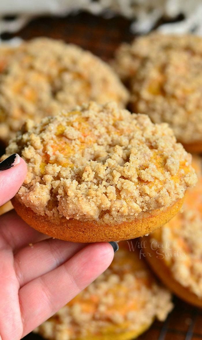 hand holding 1 pumpkin carrot cake streusel donut above a pile of stacked donuts on a wooden table