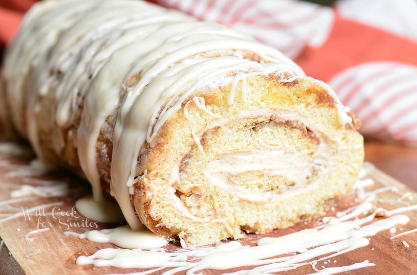 Close up view of cinnamon roll cake roll on a wooden board with icing drizzled across the top of the roll and a white and red cloth in the background