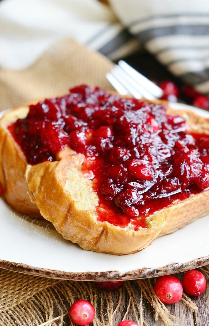 small round white plate with cranberry bliss french toast on a brown burlap placemat, a fork in the background to the right and several cranberries scattered around the plate