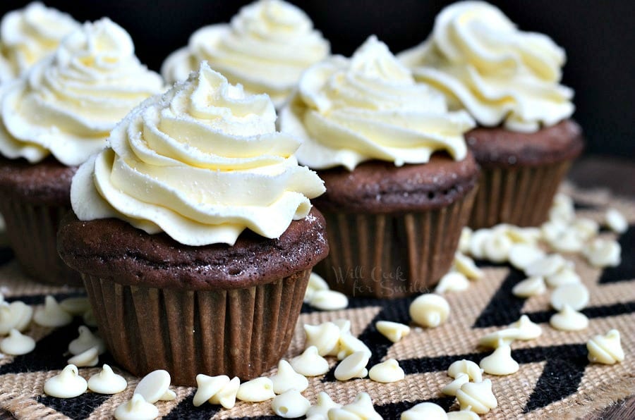 Double chocolate cupcakes with white chocolate cream cheese frosting on a white and black placemat with white chocolate chips scattered around the bottom of the cupcakes as seen close up