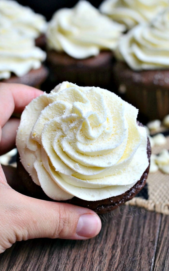 Double chocolate cupcakes with white chocolate cream cheese frosting on a white and black placemat with white chocolate chips scattered around the bottom of the cupcakes and a hand holding 1 cupcake in the foreground