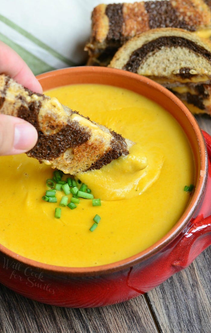 dipping grilled cheese in Pumpkin Ale Soup in a orange soup bowl with chives on top as a garnish and pumpernickel grilled cheese behind the bowl