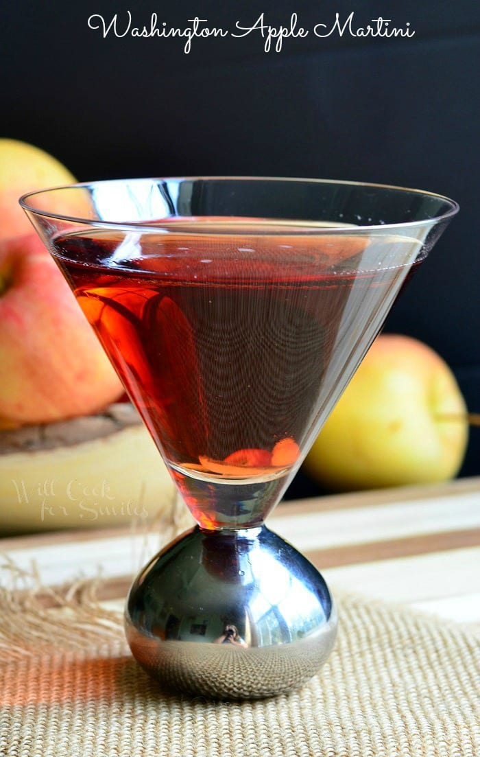 Side view of a decorative martini glass with washington apple martini on a brown placemat with a bowl of apples in the background to the left