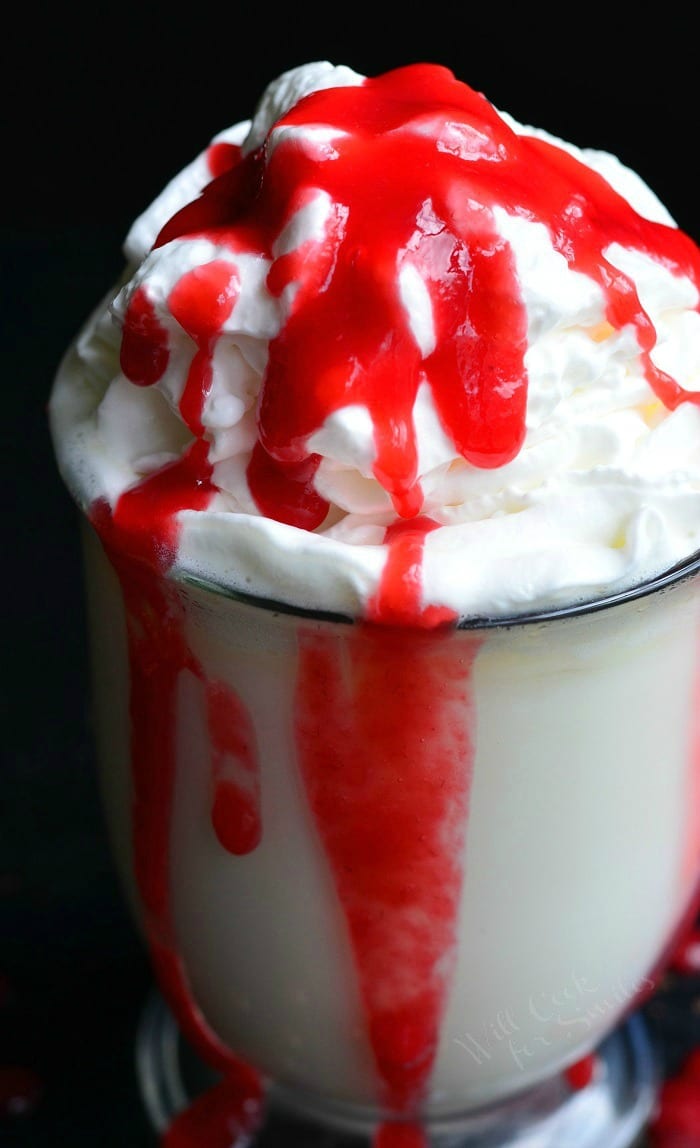 Clear glass coffee mug filled with white chocolate raspberry halloween hot chocolate on a black table with raspberry sauce splatter on table around base of glass viewed close up