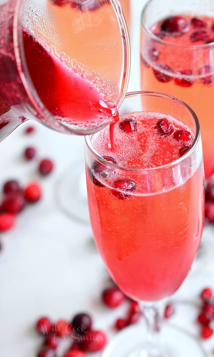 3 champagne glasses filled with cranberry pomegranate mimosas on a white table with cranberries scattered along the base of the glasses and more being poured into the glass in the foreground