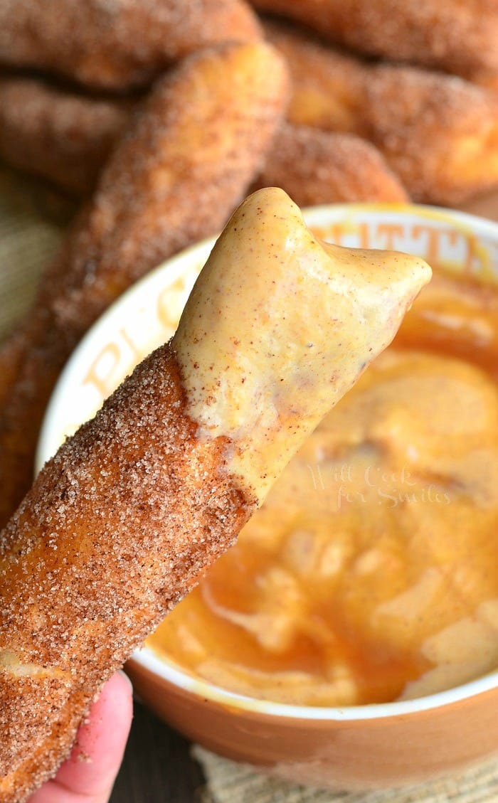 a tan bowl filled with pumpkin yogurt dip on wood cutting board with easy sugar doughnut sticks piled next to bowl and a hand holding 1 stick after being dipped in dip