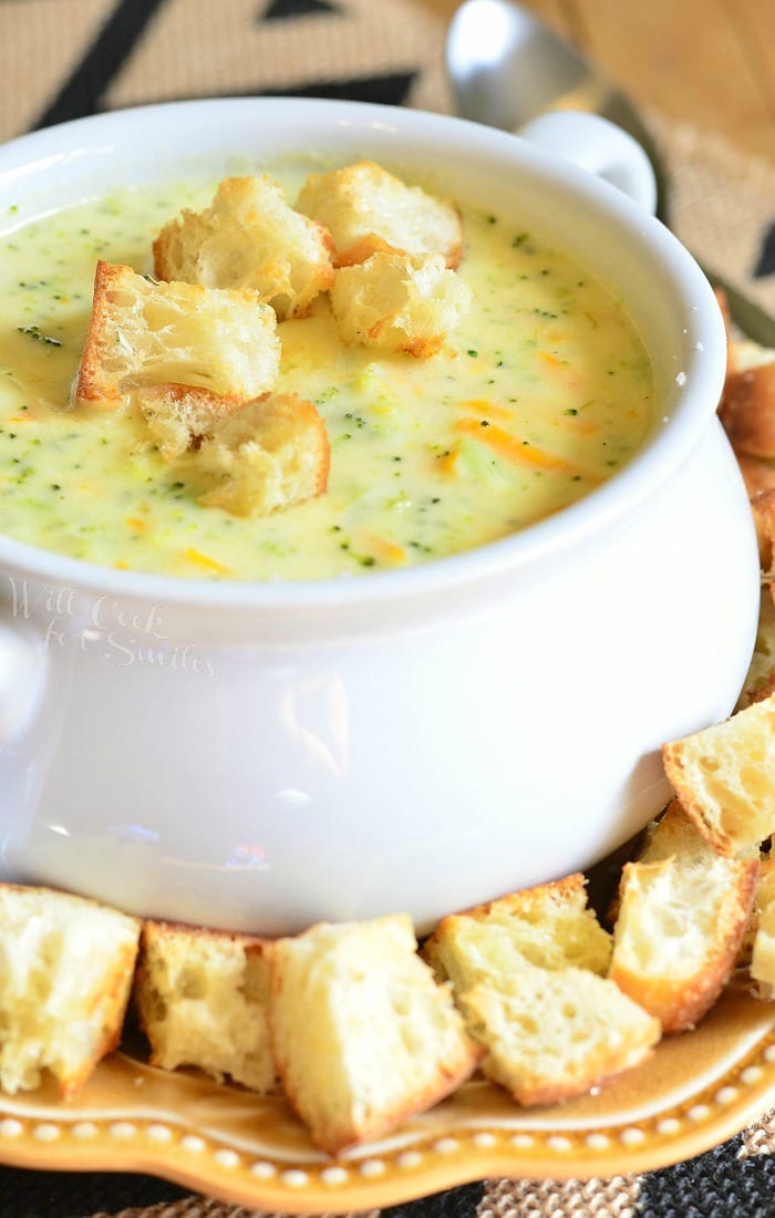 White crock filled with asiago cheese soup on a yellow plate with cut bread croutons scattered around the bowl placed on the plate with a silver spoon in the background