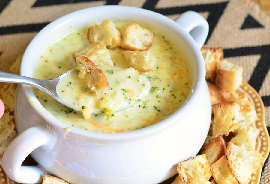 White crock filled with asiago cheese soup on a yellow plate with cut bread croutons scattered around the bowl placed on the plate viewed close up with a spoon holding one portion of soup