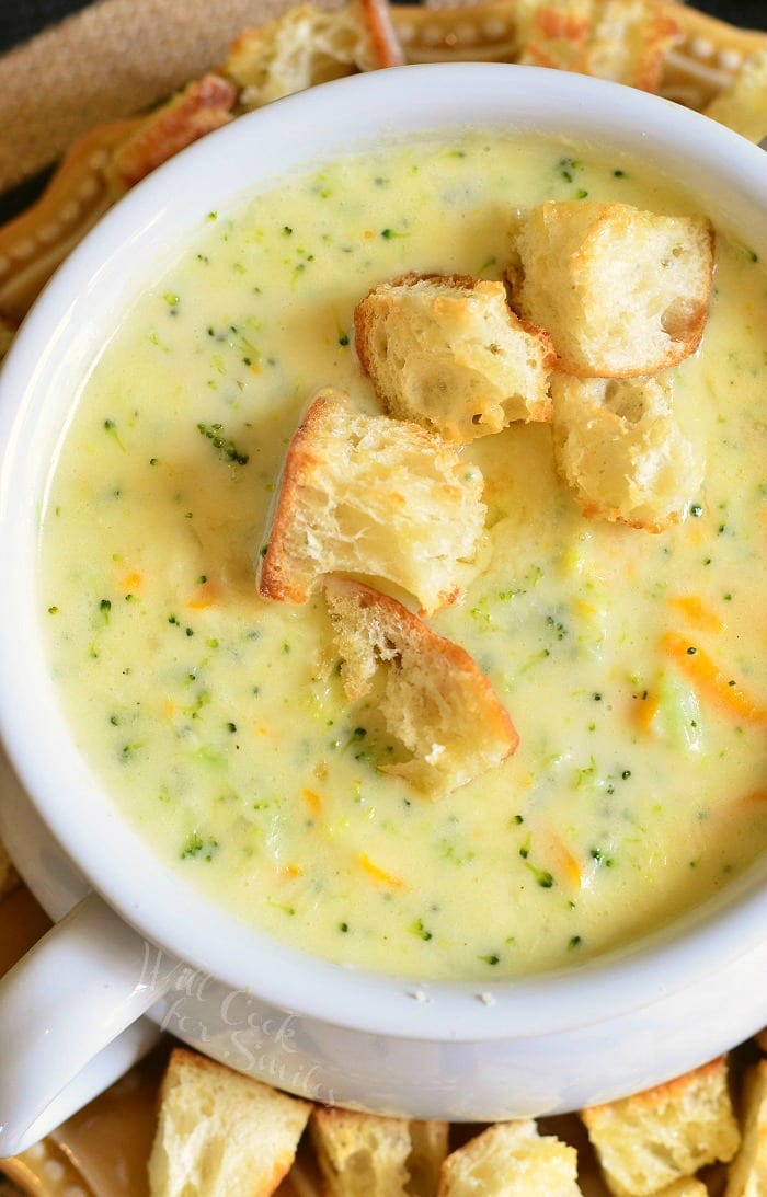 White crock filled with asiago broccoli cheese soup on a yellow plate with cut bread croutons scattered around the bowl placed on the plate as viewed from above