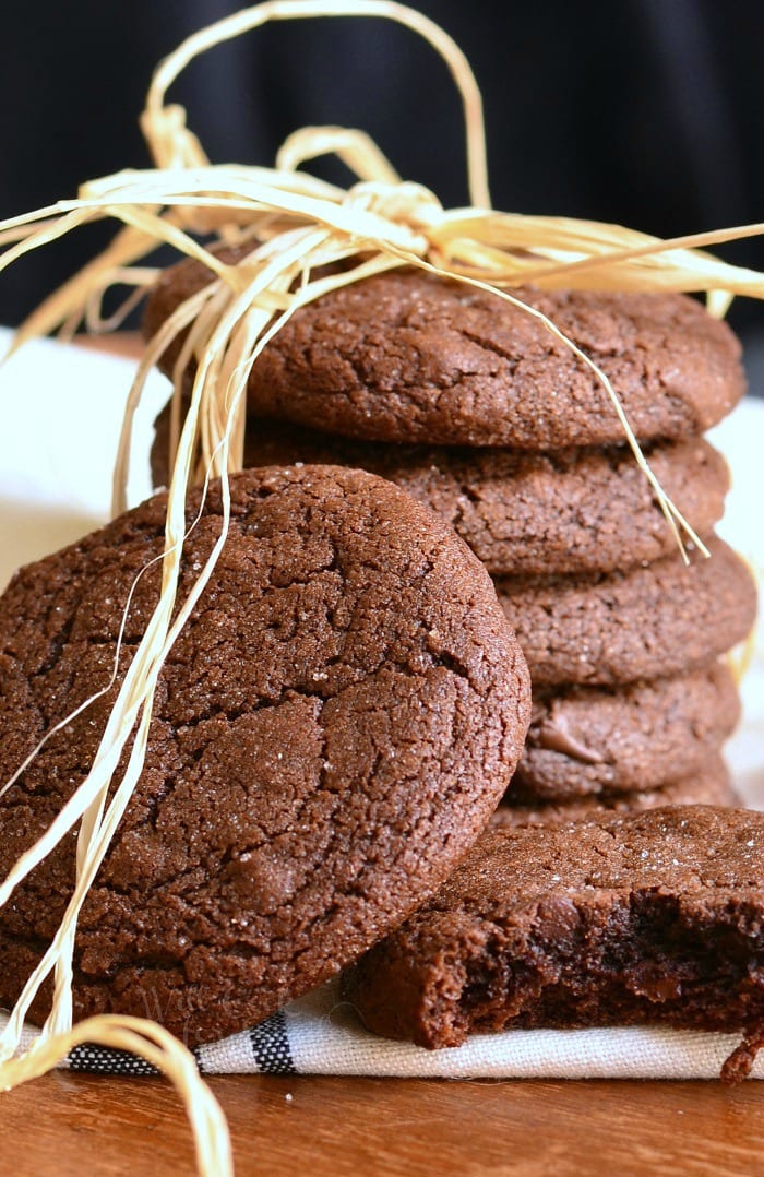 2 chocolate chocolate chip gingersnap cookies in front of additional cookies stacked behind on a white cloth while all sit on a wooden table