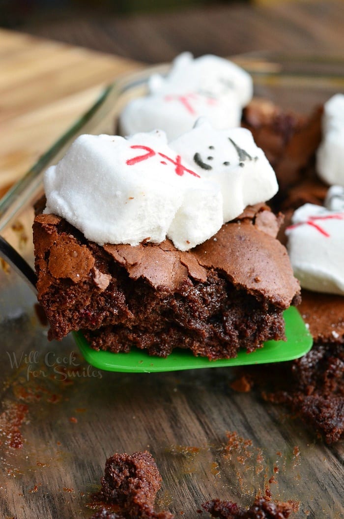 A small green spatula holding 1 gooey peppermint marshmellow brownie above a wooden table as a glass baking dish and additional brownies rest in the background
