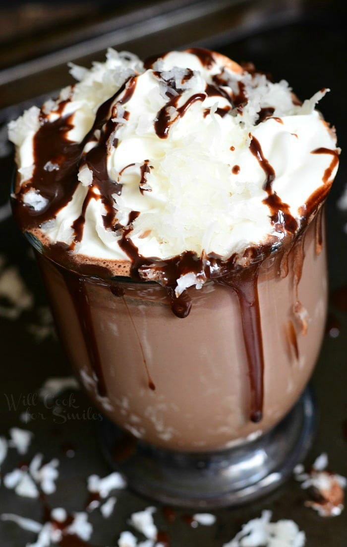 Hot Chocolate in a clear glass with whipped cream, chocolate sauce, and shredded coconut on top