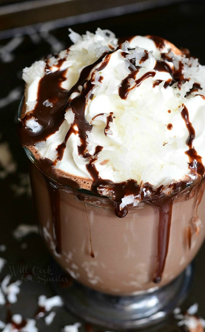 Hot Chocolate in a clear glass with whipped cream, chocolate sauce, and shredded coconut on top