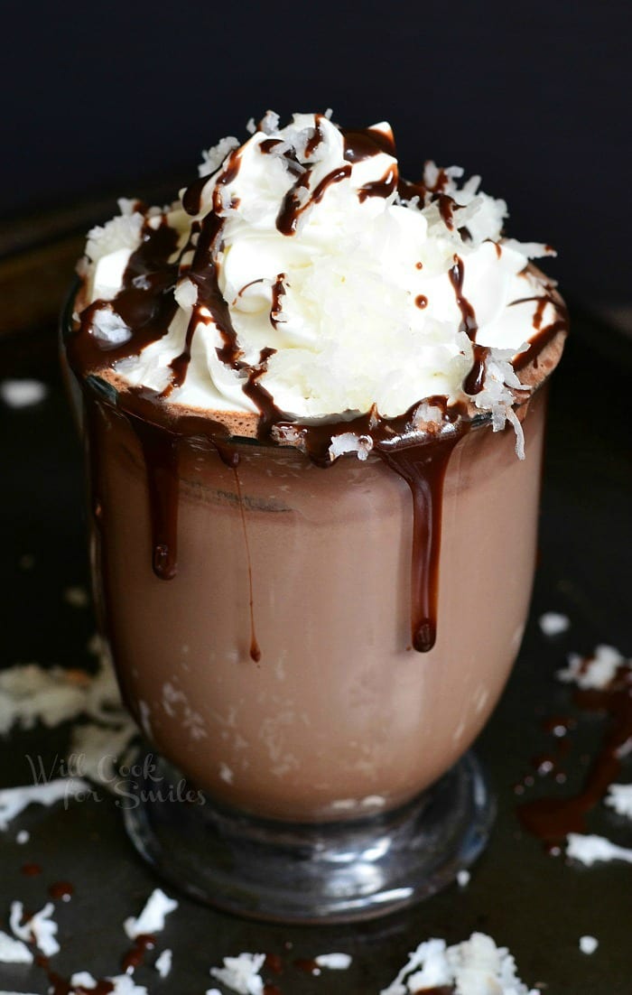 Hot Chocolate in a clear glass with whipped cream, chocolate sauce, and shredded coconut on top