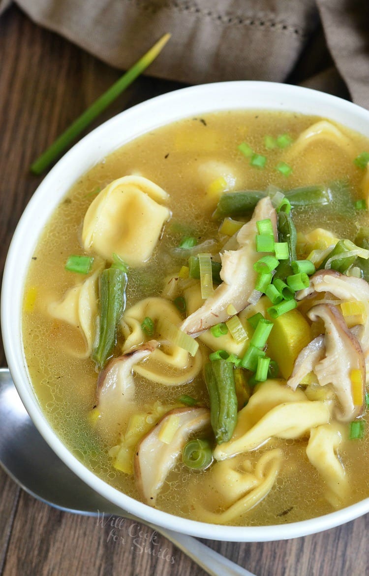 top view image Vegetable Medley Tortellini Soup in a white bowl with green onion as garnish and a spoon to the left of the bowl