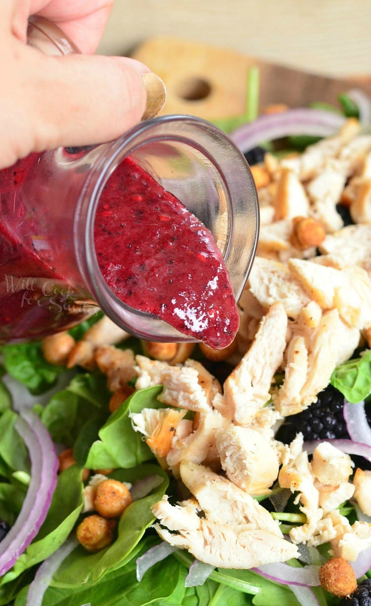 pouring salad dressing from container onto salad with lettuce, chicken, red onion, black berries and chickpeas