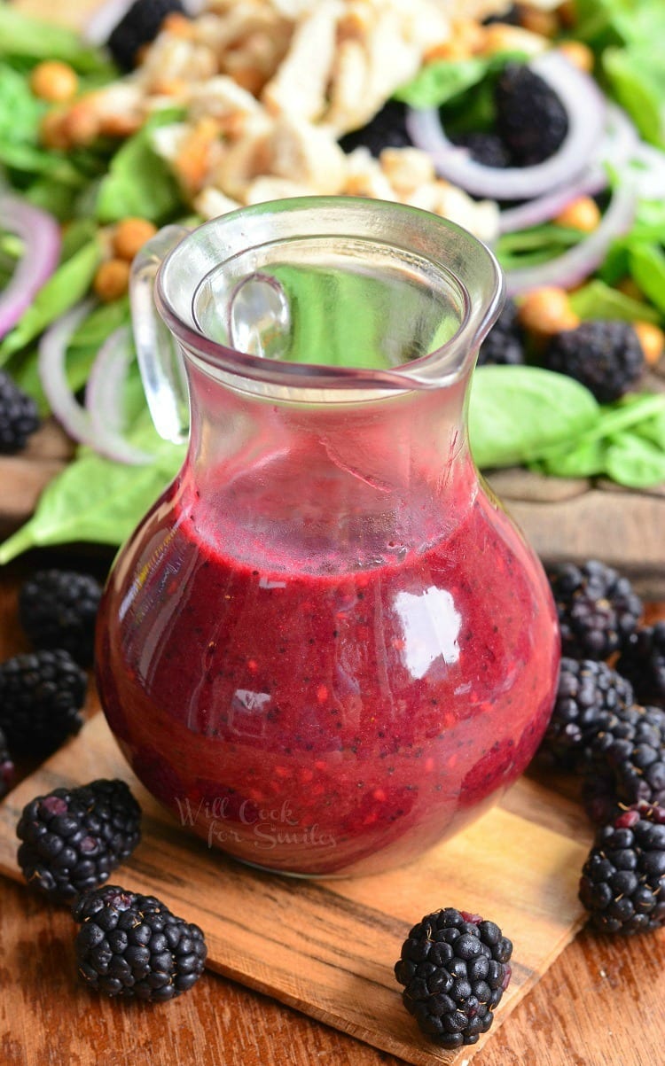 Glass of black berry poppy vinaigrette on a wooden board with blackberries scattered around the base of glass and a salad in the background