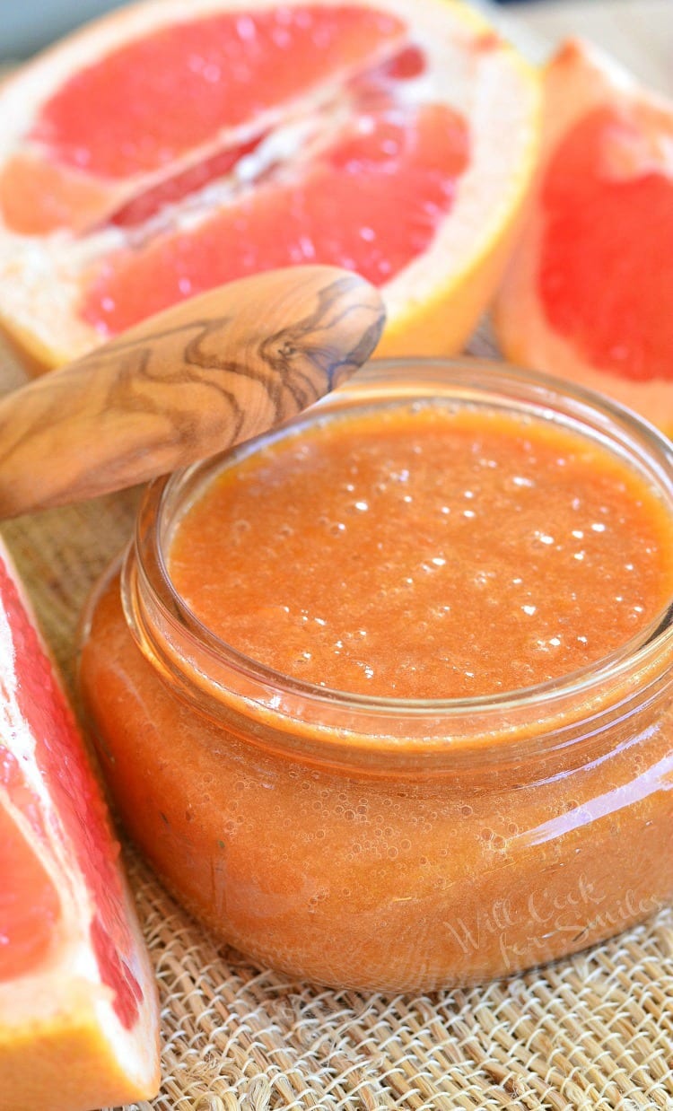 Homemade Grapefruit Sauce in a glass jar with a wood lid leaning on it and cut up grapefruit in the back ground