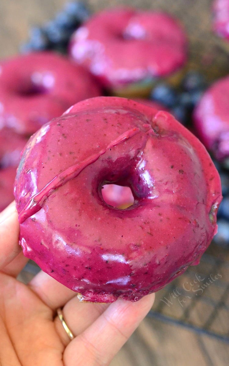 A hand holding a Blueberry doughnuts with blueberry cream whiskey glaze on a wire rack with blueberries scattered around them