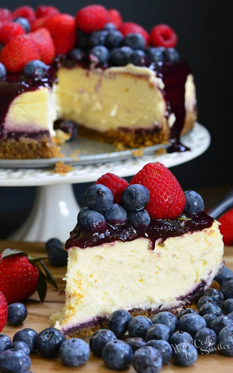 slice of cheesecake with berry sauce, raspberries, and strawberries on top on a cutting board with strawberries and blueberries around it with the rest of the cheesecake on a white cake stand in the background