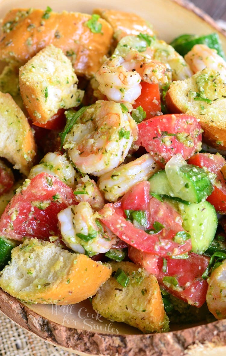 Wooden bowl filled with panzanella salad with shrimp and pesto on a tan placemat as viewed from above and close up