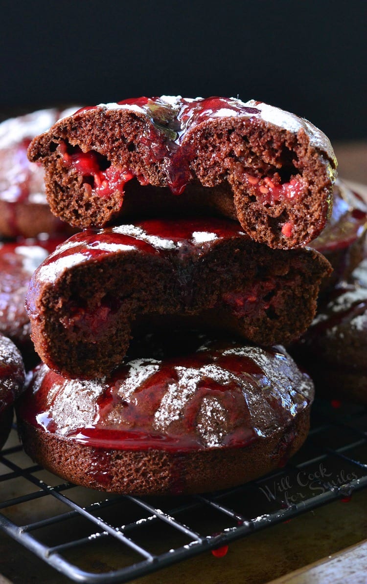 Raspberry Chocolate Doughnuts stacked up with the top one cut in half on a cooling rack with powdered sugar and raspberry sauce over the top of them on a cooling rack