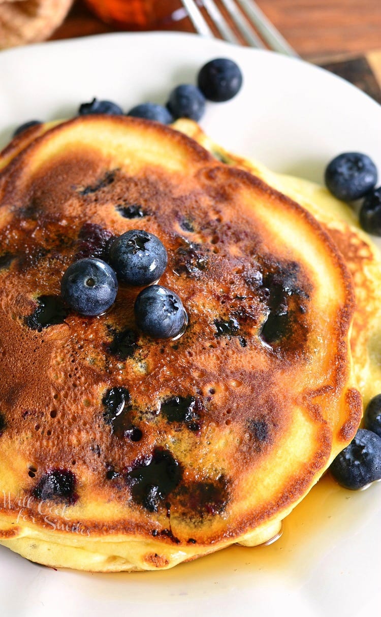 Single serving buttermilk pancakestopped with syrup and blueberries on a white plate with a fork in the background to the right.