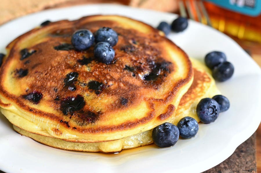 Single serving buttermilk pancakestopped with syrup and blueberries on a white plate with a fork in the background to the right as viewed close up