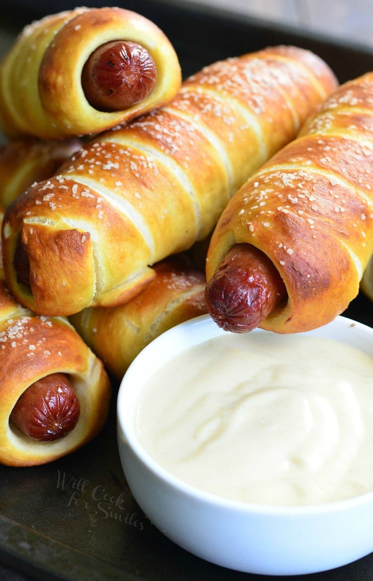 Pretzel Wrapped Hot Dogs stacked up on baking sheet with Maple Dijon Dipping Sauce in a white bowl