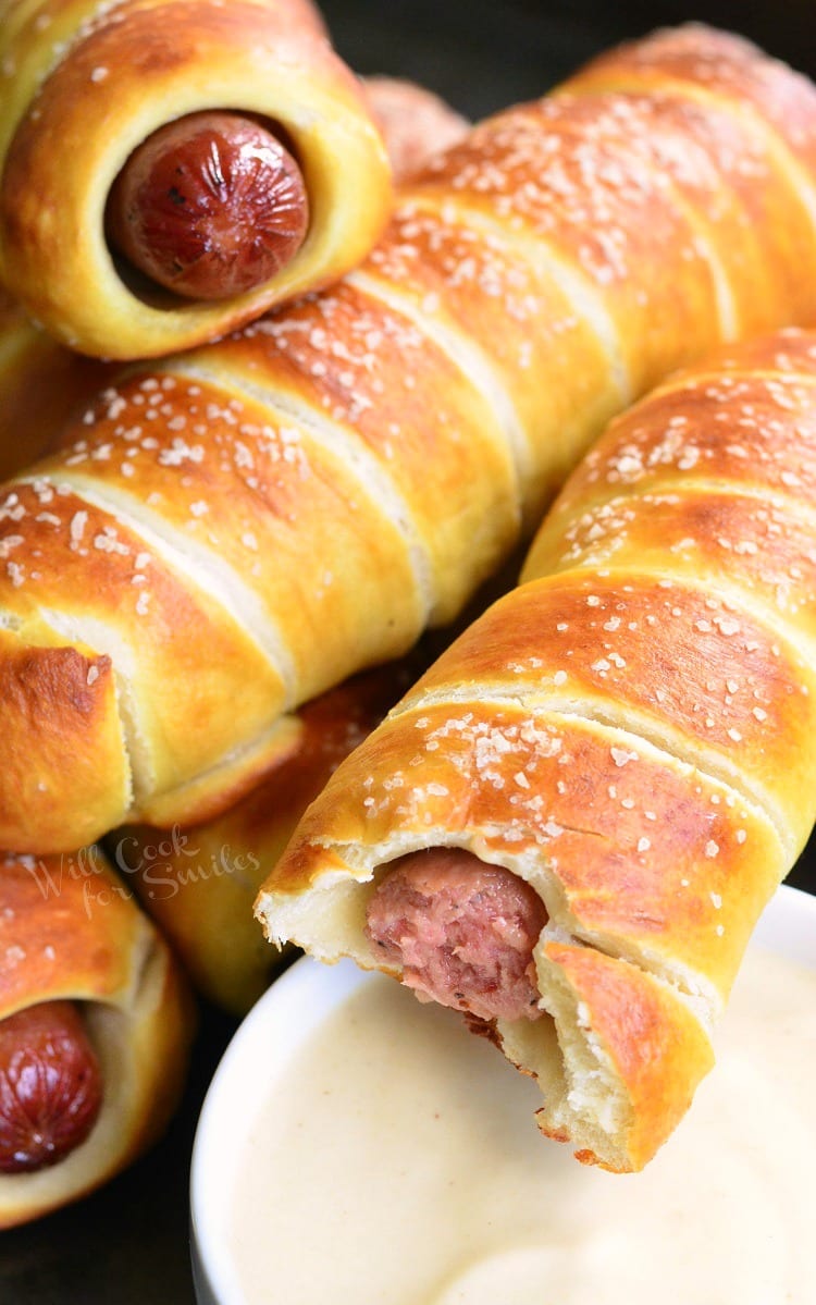 Pretzel Wrapped Hot Dogs stacked up on baking sheet with one having a bite taken from it with Maple Dijon Dipping Sauce in a white bowl