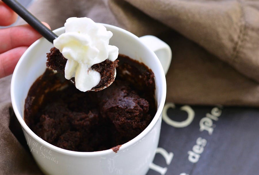 Chocolate cake in a white coffee mug with a spoon lifting some out with whip cream on top