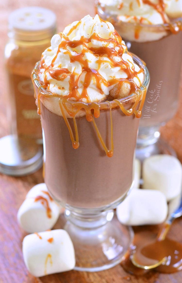 Caramel Spicy Hot Chocolate in a clear glass mug with whip cream and caramel on top on a cutting board with marshmallows around it