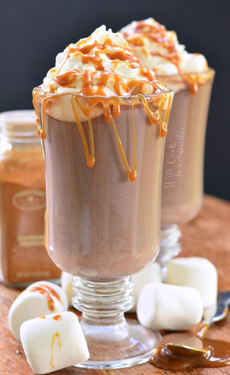 Caramel Spicy Hot Chocolate in a clear glass mug with whip cream and caramel on top on a cutting board with marshmallows around it