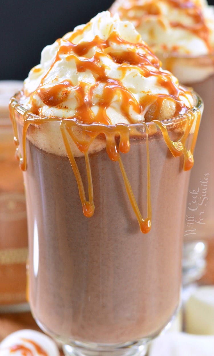 Caramel Spicy Hot Chocolate in a clear glass mug with whip cream and caramel on top on a cutting board with marshmallows around it