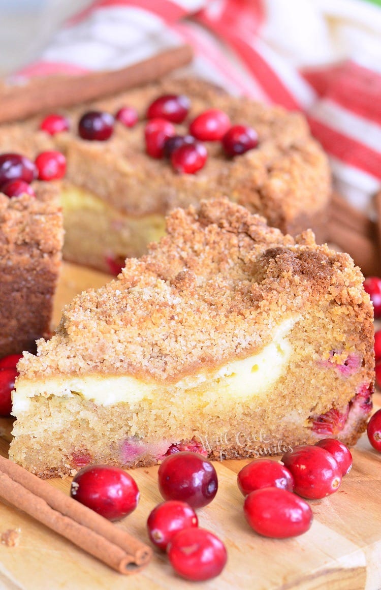 1 slice of cranberry cream cheese coffee cake on a wooden cutting board with the rest of the cake sitting behind topped with cranberries.