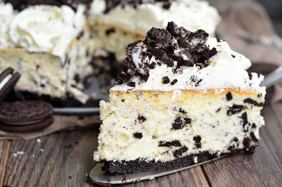 A slice of oreo cheesecake on a wood table with oreos and the rest of the cheesecake in the background