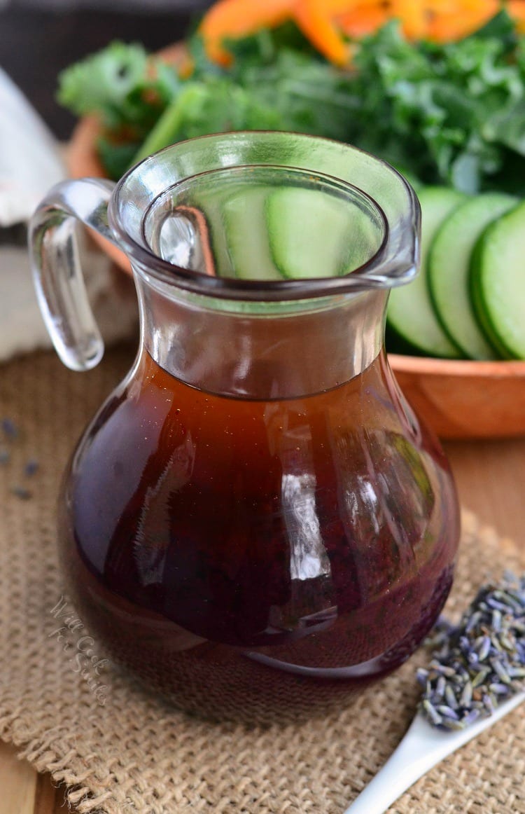Blueberry Lavender Vinaigrette in a glass container on the right is some lavender on a white spoon in the backroom is a salad in a brown bowl